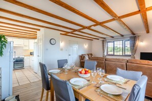 A dining area with a wooden table set for four, a kitchen in the background, and a living area with a sofa and a TV. - 3 Bed in Woolacombe (oc-s45868) (Woolacombe)