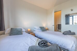 A hotel room with two beds, a small table, a lamp, a window with curtains, and a towel rack. - 3 Bed in Woolacombe (oc-s45868) (Woolacombe)