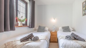 A twin room with two beds, a bedside table with a lamp, a window with curtains, and a wall-mounted picture frame.