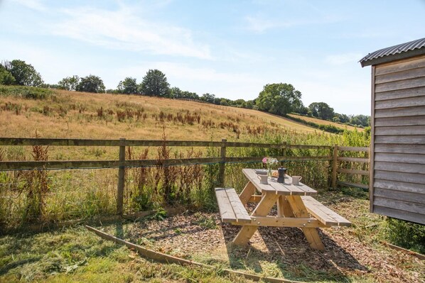 A picnic table with benches in a grassy area with a wooden fence and a small building in the background.