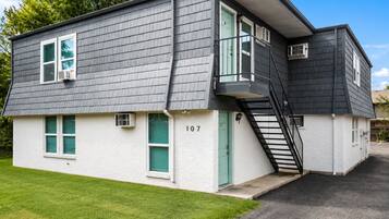 Un bâtiment à deux étages, à l’extérieur gris, avec une porte verte et un escalier apparent menant à l’étage supérieur.