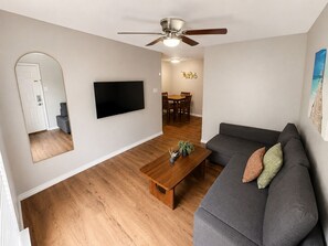 Een moderne woonkamer met een flatscreen televisie, een plafondventilator, een houten salontafel en een grijze bank met gestreepte kussens.