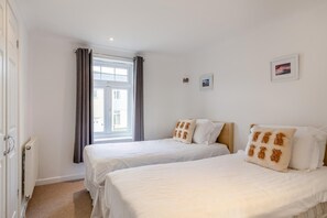 A hotel room with two beds, a window with curtains, a radiator, and a small picture on the wall. - 4 Bed in Newquay (oc-a45833) (Black Cross)