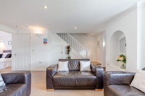 A spacious living room with a leather sofa, a staircase, and a white wall with a window. - 4 Bed in Newquay (oc-a45833) (Black Cross)
