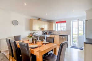 A modern kitchen with wooden cabinets, a dining table with chairs, and a clock on the wall. - 4 Bed in Newquay (oc-a45833) (Black Cross)