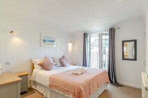 Ein Schlafzimmer mit einem Bett, Nachttischen, einem Fenster mit Vorhängen, einem gerahmten Bild und einer Wandleuchte.