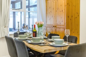 A dining table set with plates, glasses, and a bottle of wine, with a vase of flowers and a window in the background. - 3 Bed in Brancaster (oc-m45869) (Brancaster)