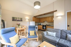 Ein Wohnzimmer mit einer Couch, einem Sessel und einem Esstisch mit Stühlen. Eine Küchenzeile mit Holzschränken und Geräten ist ebenfalls vorhanden.