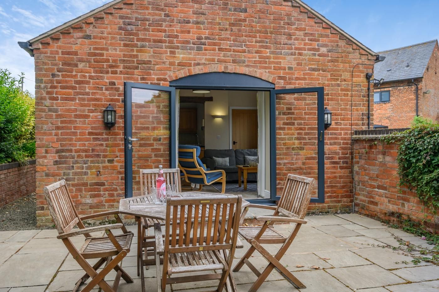 A brick building with a patio area featuring a table and four chairs, a sliding glass door leading to an interior space with a sofa and a chair.