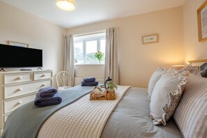 Ein Schlafzimmer mit einem Bett, einem Fernseher auf einer Kommode, einem Fenster mit Vorhängen und einem Nachttisch mit einer Tablett voller Gegenstände.