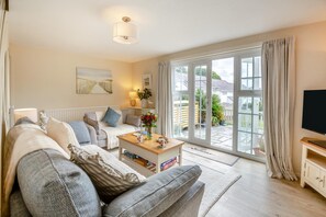 Ein Wohnzimmer mit einer Couch, einem Couchtisch und einem Fernseher. Ein großes Fenster mit Vorhängen und eine Tür, die nach draußen führt.