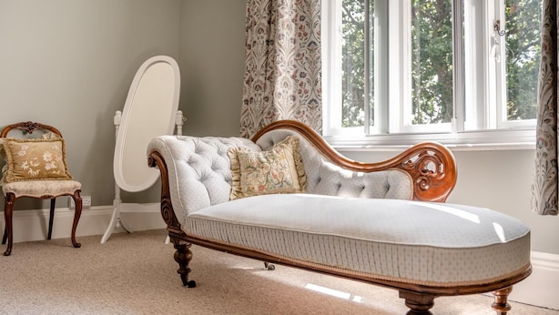 A room with a large, tufted sofa, a small wooden chair, a full-length mirror, and a window with curtains. - 3 Bed in Poole (oc-v45688) (Sandbanks)