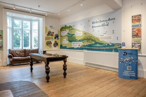 A room with a large wall mural titled 'Waters Journey Across Brownsea Island,' a brown leather sofa, a wooden table, and a blue informational stand. - 3 Bed in Poole (oc-v45688) (Sandbanks)