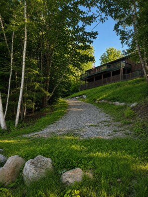 A gravel path leading to a wooden cabin with a balcony, surrounded by trees and grass. - Antler Lake Haus - Private Waterfront & ATV/Snowmobile Trail Access (Bigfork)