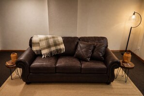 A brown leather sofa with a plaid throw blanket, two side tables with lamps, and a herringbone-patterned rug.