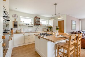 A modern kitchen with white cabinetry, a central island, and a dining area with wooden chairs. - 3 Bed in Braunton (oc-m45420) (Braunton)