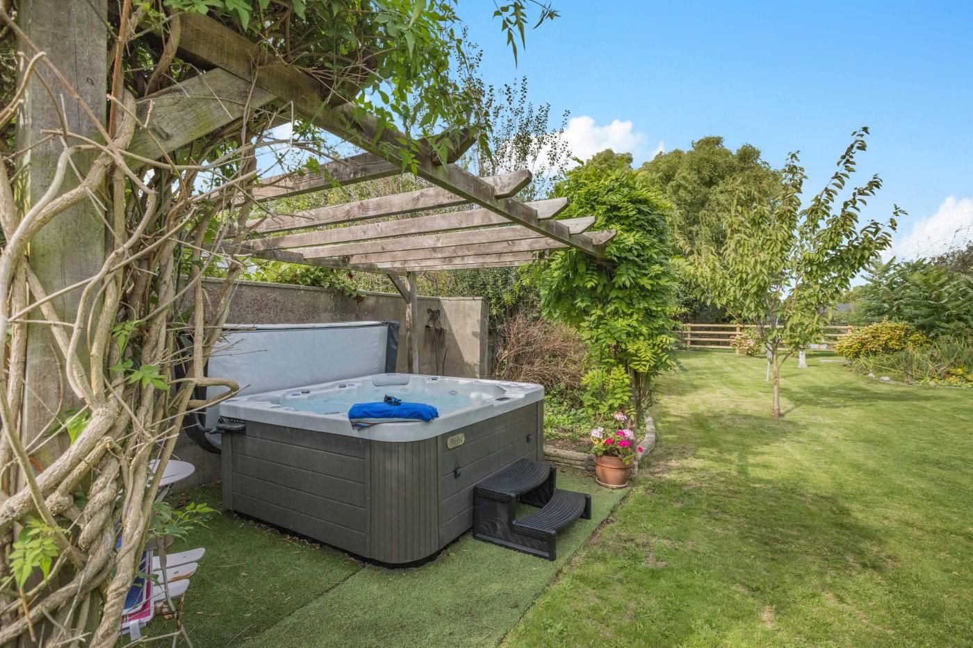 Un jacuzzi bajo una pérgola de madera en un jardín exuberante con una valla de madera y árboles al fondo.