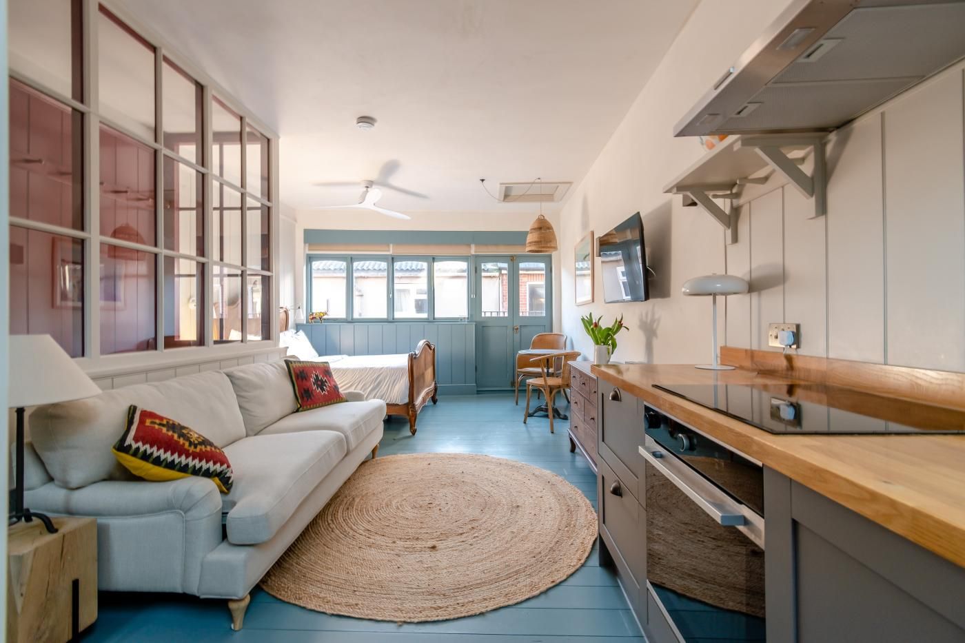 A compact living space with a kitchenette, a sofa with a patterned cushion, a dining area with a table and chairs, and a large window with a view.