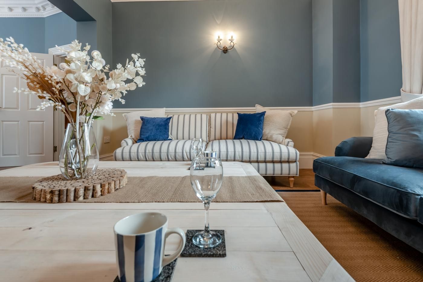 Ein Wohnzimmer mit einem gestreiften Sofa, einem blauen Sitzsofa, einer Vase mit weißen Blumen, einem Weinglas und einer Kaffeetasse auf einem Untersetzer.