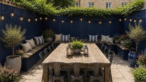 An outdoor dining area with a wooden table and chairs, surrounded by planters and a blue fence with climbing plants. - 9 Bed in Tenby (oc-h45564) (Tenby)