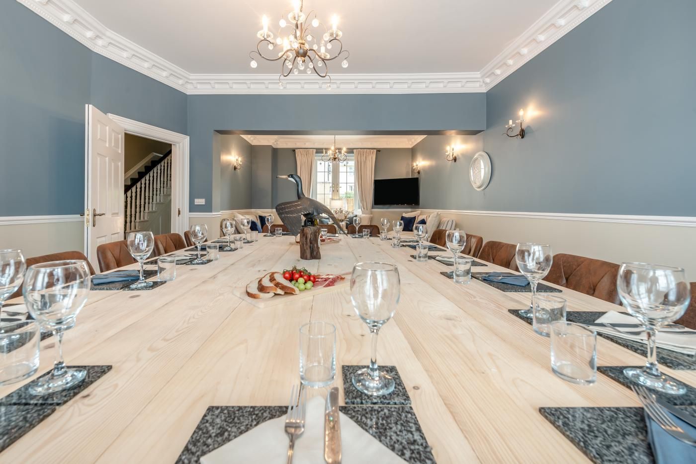 A dining room with a long table set for a meal, a chandelier, and a television in the background.