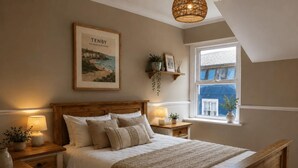 A bedroom with a wooden bed, bedside tables with lamps, a framed picture on the wall, and a window with a view. - 9 Bed in Tenby (oc-h45564) (Tenby)