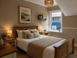 A bedroom with a wooden bed, bedside tables with lamps, a framed picture on the wall, and a window with a view. - 9 Bed in Tenby (oc-h45564) (Tenby)