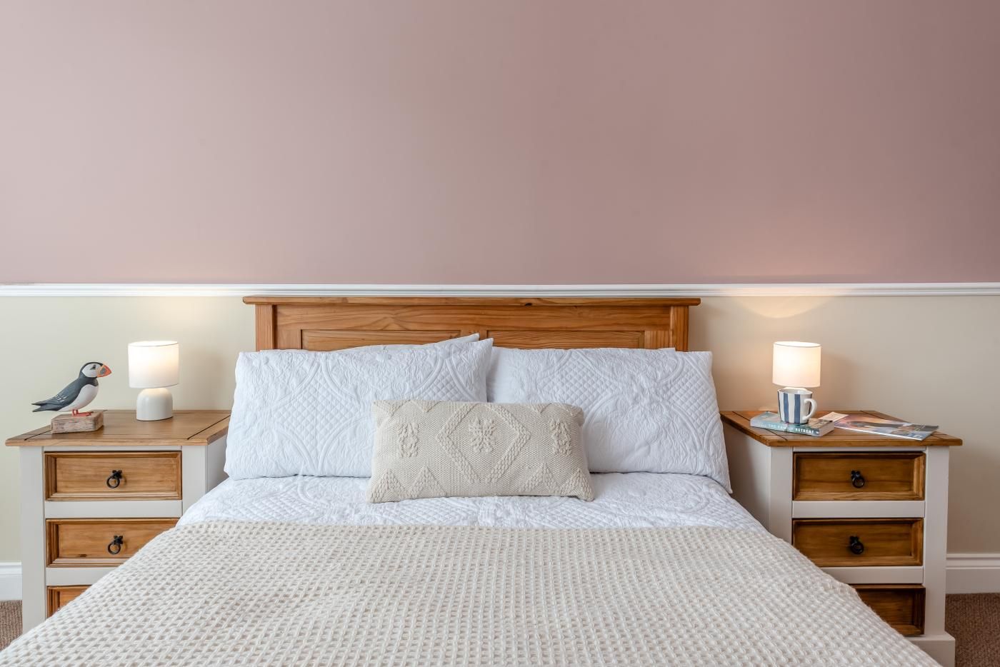 Una cama con cabecera de madera, dos mesitas de noche con lámparas, y un pájaro decorativo sobre una de ellas.