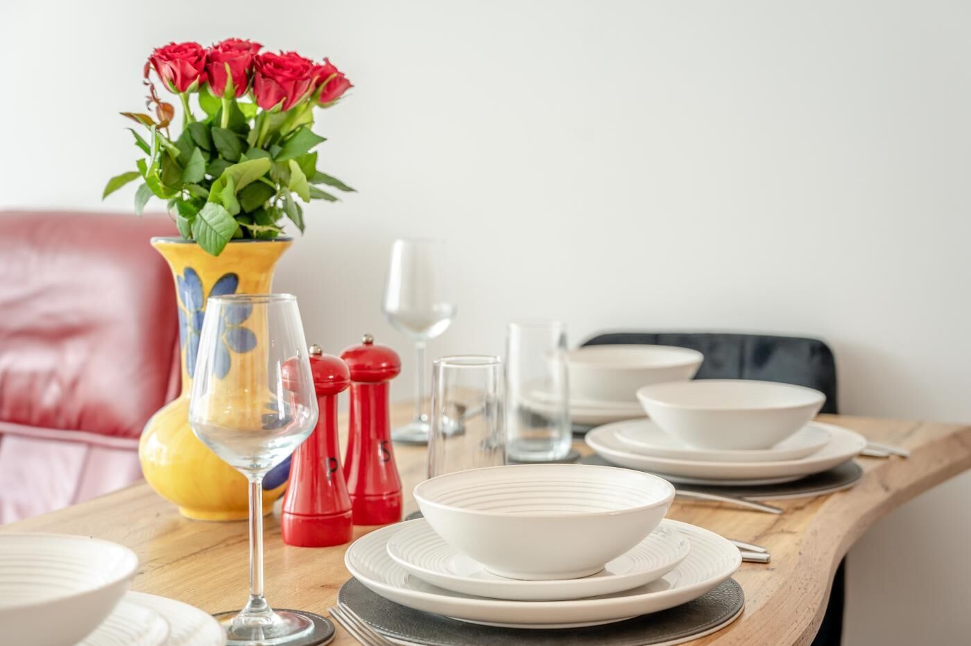 A dining table set with white plates, bowls, and a wine glass, accompanied by red salt and pepper shakers, and a vase with red roses.