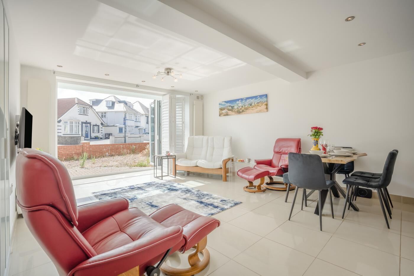 A spacious living area with a red leather sofa, a dining table with chairs, and a large window offering a view of the neighborhood.