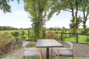 Un área de estar al aire libre con una mesa y dos sillas, rodeada de un cerramiento y árboles.