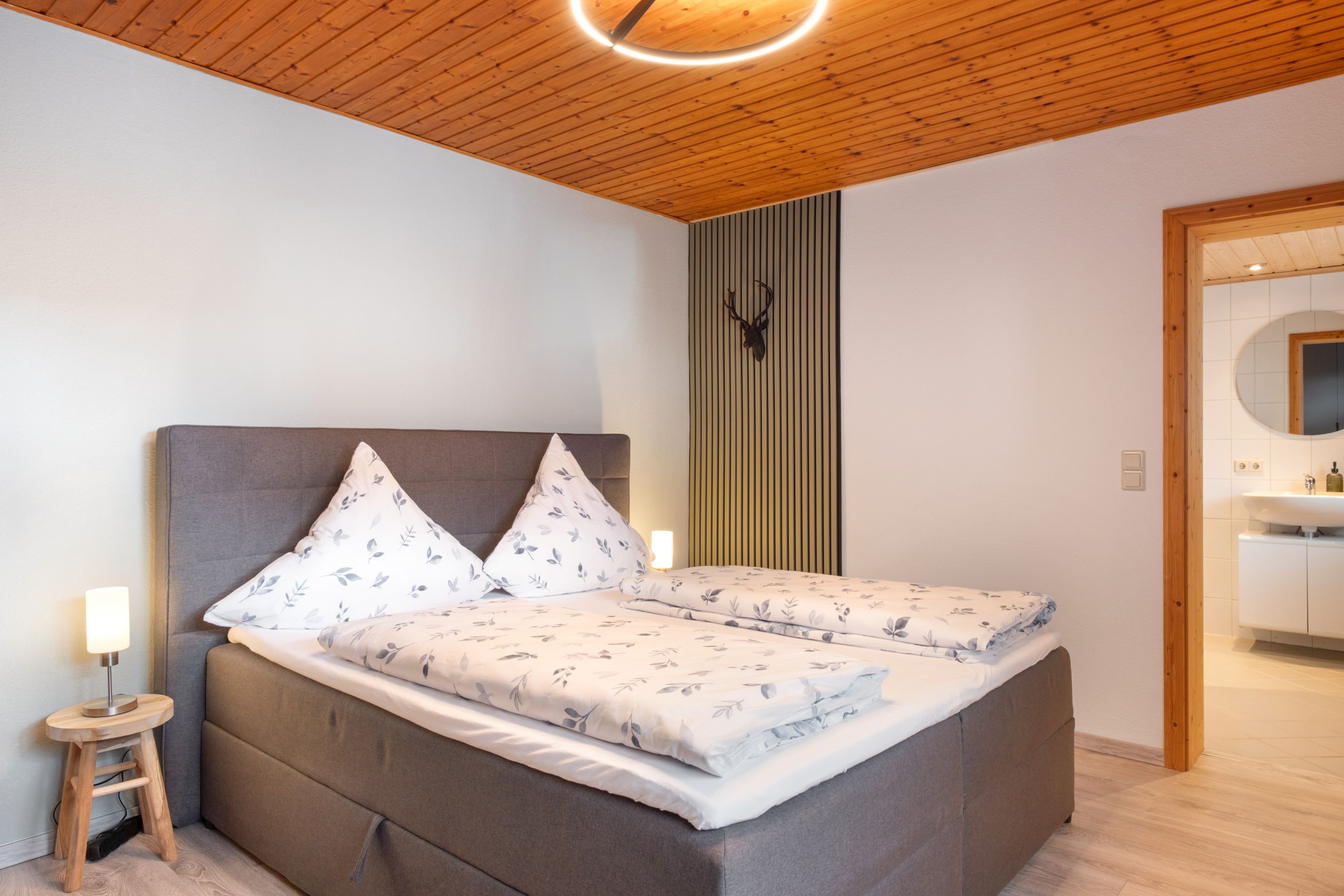 A bedroom with a bed, pillows, a bedside table, a wall-mounted light, and a wooden ceiling.