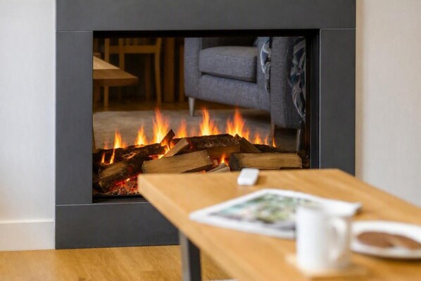 A modern fireplace with burning logs, a wooden table with a white mug, and a grey sofa in the background.