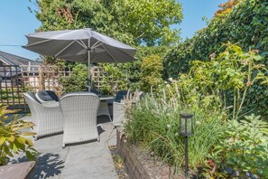 An outdoor patio with wicker furniture and a large umbrella, surrounded by lush greenery and a stone pathway.
