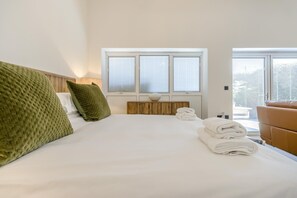 A hotel room with a large bed, a wooden bedside table, a window with blinds, and a view of the outdoors.