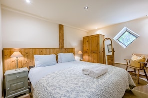 Quarto com cabeceira de madeira, cama com colcha floral, mesa de cabeceira com abajur, guarda-roupa de madeira, espelho e janela com vista para o verde.
