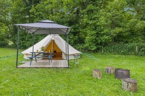 Uma tenda com uma mesa de piquenique e uma pequena mesa por perto, em meio a uma área gramada e árvores.