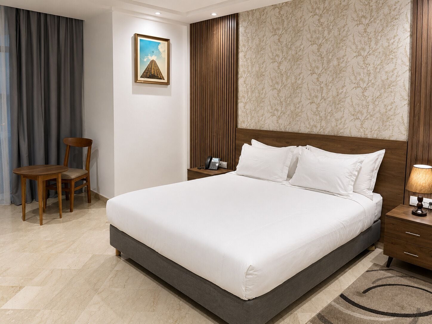 Una habitación de hotel con una cama grande, una mesita de noche de madera, una silla, un escritorio y un cuadro en la pared.