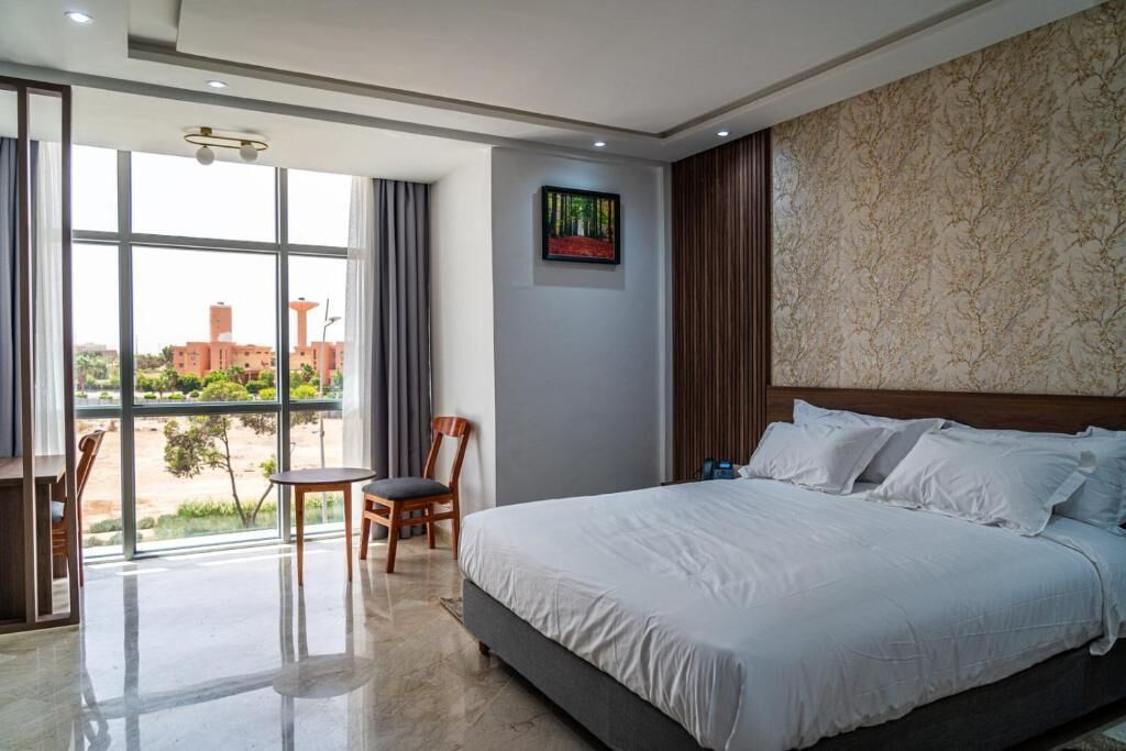 Une chambre d’hôtel avec un grand lit, une chaise, une table et une vue sur le paysage urbain grâce à de grandes fenêtres.