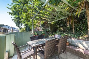 Um pátio externo com mesa e cadeiras, cercado por vegetação e uma estrutura de pérgola.