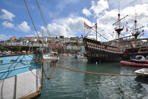 Um porto com um grande navio à vela atracado, rodeado por barcos menores e edifícios ao fundo.