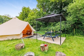 Une tente avec une table de pique-nique attenante et une balançoire installée dans un espace verdoyant bordé d’arbres.