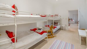 Quarto com beliche, roupa de cama branca e almofadas vermelhas, uma mesa redonda pequena e um tapete listrado.