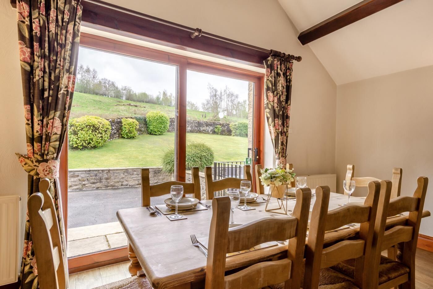 A dining area with a table set for four, wooden chairs, and a view of a green landscape through large windows.