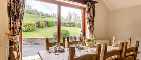 A dining area with a table set for four, wooden chairs, and a view of a green landscape through large windows.