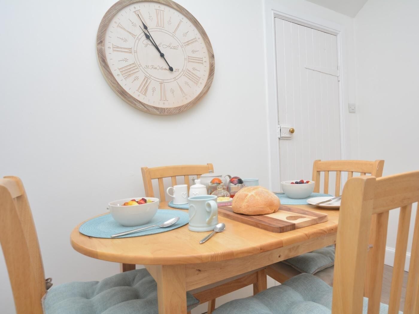 Een eetgedeelte met een houten tafel, stoelen, een klok en een deur op de achtergrond.