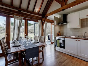 A dining area with a wooden table set for four, a kitchen with white cabinets and a stove, and a view of the outdoors through large windows. - 2 Bed in Brighton (oc-80378) (Poynings)