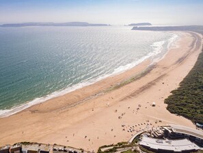 Ein Luftbild zeigt einen Strand mit einer gewundenen Promenade, einer Sandküste und ein großes Gebäude mit flachem Dach.
