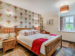 Ein Schlafzimmer mit einem hölzernen Bett, einem Nachttisch, einer Lampe, einem Fenster mit Vorhängen und einer Wand mit einem Blumenmuster.