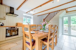 Ein Essbereich mit einem Holztisch, gedeckt für vier Personen, umgeben von Holzstühlen. Eine Küche mit einem eingebauten Backofen und einem Fenster über der Spüle ist sichtbar.